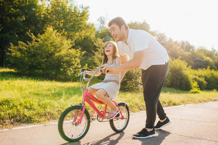 Happy father rejoices that her daughter learned to ride a bike, Father holding his daughter while she is riding bike, outdoors, the streetの写真素材