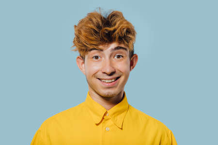 Happy young male with stylish hairdo looking at camera and smiling friendly while standing against blue backgroundの写真素材
