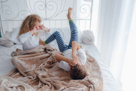 High angle of little sister and brother in pajamas awakening in cozy bed in light bedroom in morningの写真素材