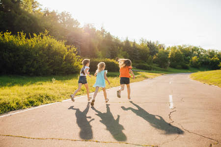Back view of group of cheerful little girls having fun and running together on asphalt road while enjoying summer day in natureの写真素材