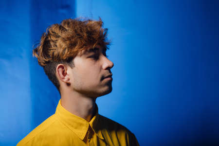 Serious millennial male with trendy dyed haircut wearing casual yellow shirt looking away against blue backgroundの写真素材