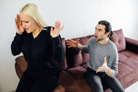 Aggressive Caucasian Husband Shouting At Unhappy Victimized Wife Sitting On Couch At Home. Family Conflicts, Quarrels And Domestic Violence, Marriage And Relationship Crisis Concept.の写真素材