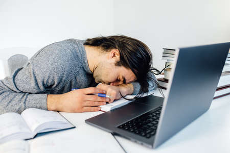 tired businessman sleeping while calculating expenses at desk in office. Young caucasian male employee take a nap in the office on a table with tablet and laptop computer.の写真素材