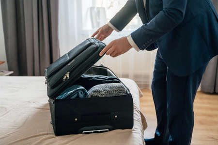 Unrecognizable male entrepreneur opening suitcase and putting garments on bed while unpacking luggage in hotel room during business tripの写真素材