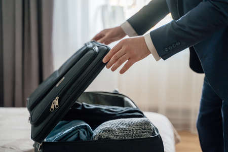 Unrecognizable male entrepreneur opening suitcase and putting garments on bed while unpacking luggage in hotel room during business tripの写真素材