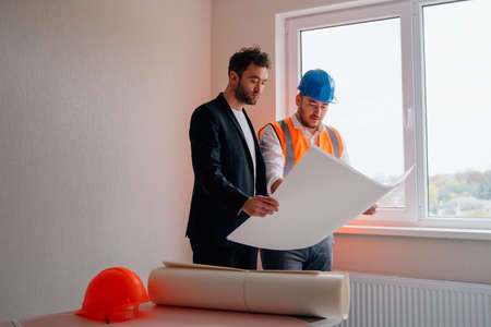 Two men Architect and engineer in vests and hardhats inspecting and discussing blueprint while working in finished room of apartment buildingの写真素材