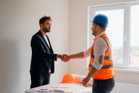 Side view of businessman and male contractor talking and shaking hands over table while working on blueprint in apartmentの写真素材