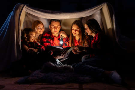 happy family mother father and children reading a book with a flashlight in a tent at home. Dad reads the story at night in the tent for the whole familyの写真素材
