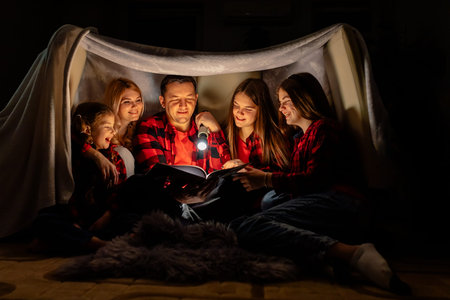 family, hygge and people concept - happy mother, father and daughters reading book with torch light...