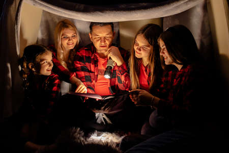 Caucasian family. Dad reads the story sitting in a teepee, reading stories with the flashlight in dark room. Home comfort, family, love, storytelling time.の写真素材