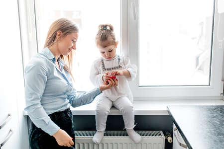 Young caucasian mother and her cute curly toddler daughter. they have fun together at home in the kitchen by the windowの写真素材