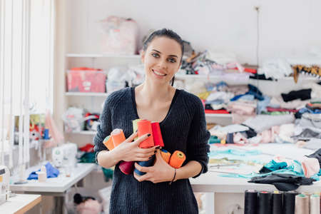 cute caucasian Seamstress or dressmaker keeps colorful spools of thread in hands on atelier shop background, front view.の写真素材