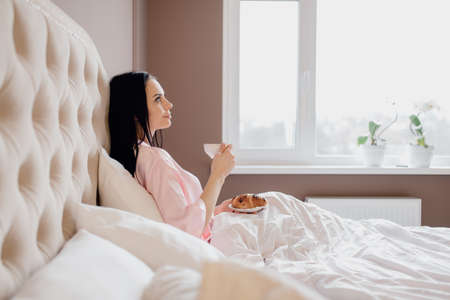 side view of the beautiful brunette girl serving coffee in the morning in her bed, at home. caucasian woman smiles a gorgeous morningの写真素材