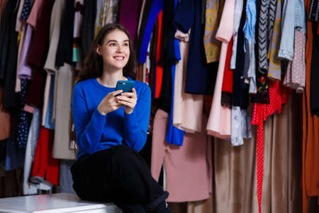 happy beautiful teen blogger writes a text on the phone. Beautiful girl front of modern clothes wardrobe closet in the shopping store. sale, consumerism, fashion, communication and people conceptの写真素材