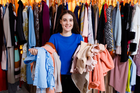 happy nice caucasian teenage girl with clothes on her arms in the clothing store. he doesn't know what to choose. looking at camera. Beautiful young woman near rack near hangers with clothing. shopping store, clothing storeの写真素材