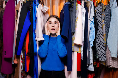 Perplexed beautiful Caucasian teenage girl in clothing store. he doesn't know what to choose. looking at camera. Beautiful young woman near rack near hangers with clothing. shopping store, clothing storeの写真素材