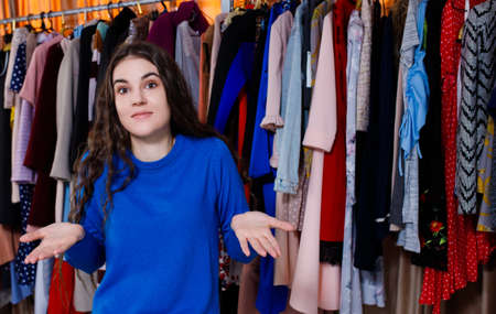 confused girl does not know what to choose, looking at camera. Beautiful young woman near rack near hangers with clothing. shopping store, clothing storeの写真素材