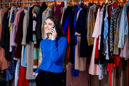 smiling beautiful brunette teenager dressed in blue sweater talking on the phone, Beautiful girl front of modern clothes wardrobe closet in the shopping store. shopping, fashion, style and people conceptの写真素材