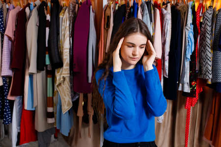 teenager dressed in a blue sweater with her hands on her head, has a headache, the choice problem, is in the clothing store. Beautiful young woman near rack near hangers with clothing. shopping store, clothing storeの写真素材