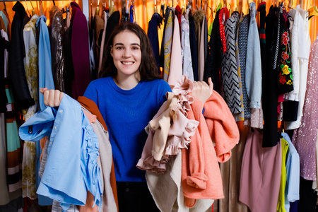 happy nice caucasian teenage girl with clothes on her arms in the clothing store. he doesn't know what to choose. looking at camera. Beautiful young woman near rack near hangers with clothing. shopping store, clothing storeの写真素材