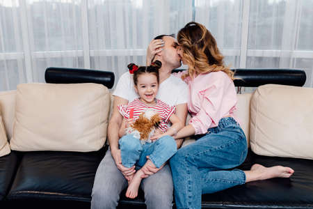 Enjoying time together. Young loving father carrying his smiling daughter and kissing his wife while spending free at home on sofa.の写真素材