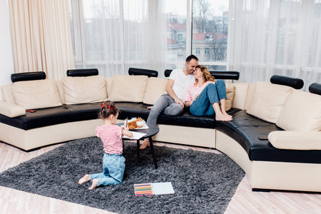 little children girl sitting on floor pencil drawing on paper. Couple sitting on sofa and little daughter drawing, daughter have a fun on warm carpet floor with colorful pencils.の写真素材