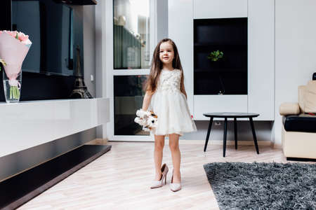 Cute little fashionista in white dress. Happy child girl try on outfits and mom's shoes in living room. smiling funny little baby girl walks at home trying mom's high heel shoesの写真素材