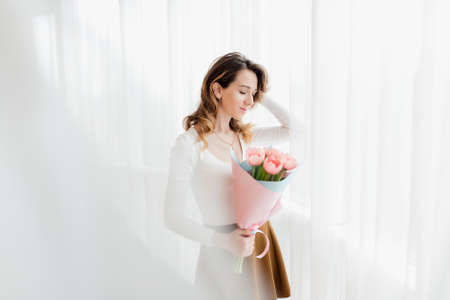 Beautiful young brunette woman with tulip bouquet. Spring portrait. At home interior. Happy international women's day! Celebrating 8th of March.の写真素材
