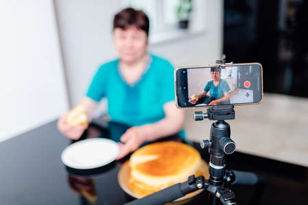 Senior woman blogger sit at kitchen at home while being recorded on smartphone on stabilizer for culinary blog. grandmother recording vlog for social blogger. selective focusの写真素材