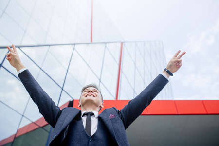 Happy overjoyed caucasian Businessman celebrate good news surprise at at work with modern office building, excited man winner screaming yes rejoicing successの写真素材