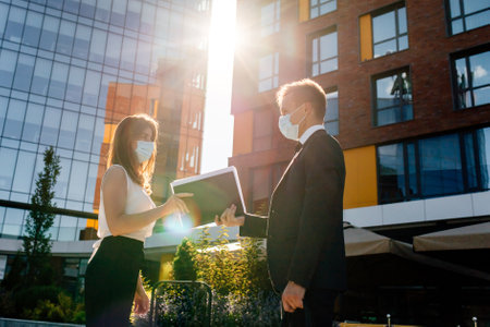 Low angle side view of serious young business partners in formal clothes and medical masks share documents while standing near modern office building in city downtownの写真素材