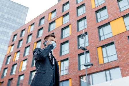 Low angle of serious young male entrepreneur in formal suit and medical mask for virus prevention talking on smartphone while standing in city downtownの写真素材