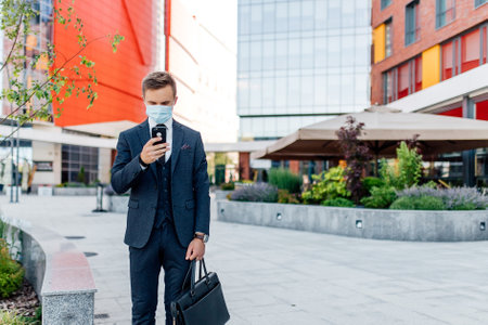 Serious well dressed young businessman in formal suit and medical mask messaging on mobile phone while standing on city street near contemporary buildingsの写真素材