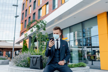 businessman in formal suit and medical mask messaging on smartphone while standing on city street near contemporary buildingsの写真素材
