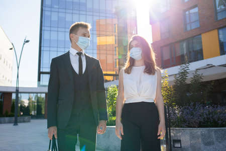 Low angle of cheerful young man and woman in elegant formal outfits and protective masks talking and smiling while having meeting near contemporary office building in city downtownの写真素材