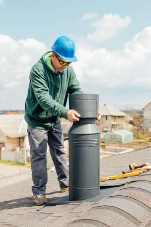 male worker with blue helmet installs an iron chimney. worker with sunglasses repairing chimney on roof.の写真素材
