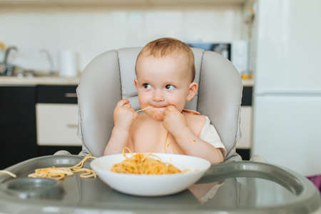 little Boy eating spaghetti look hungry and pasta sauce messes all around his face while having breakfast. kid boy eats pasta with sauce sitting in kitchenの写真素材