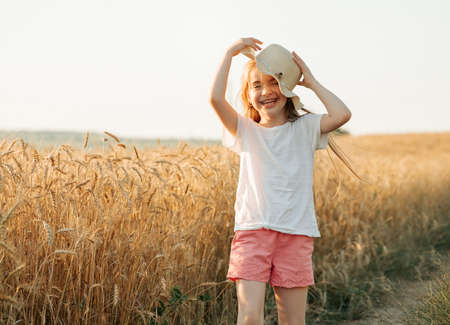 kid girl with hat, happy smiles beautifully while in golden wheat field. childhood in the countryside in the village, beautiful child smiles at cameraの写真素材