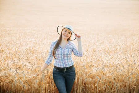 Young caucasian smile woman farmer looking at camera and wear hat and dress in squares stand in wheat field. Farm female Worker Working in wheat Fields.の写真素材