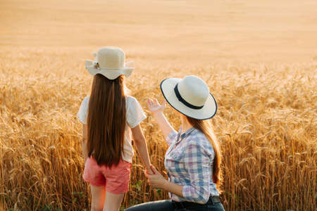 rear view of mother and daughter pointing to wheat field. Mom and her kid family of farmers with hat in golden wheat field, harvest time.の写真素材