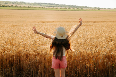rear view of little kid girl with hat raise your hands up in wheat land. happy little girl jumps up...
