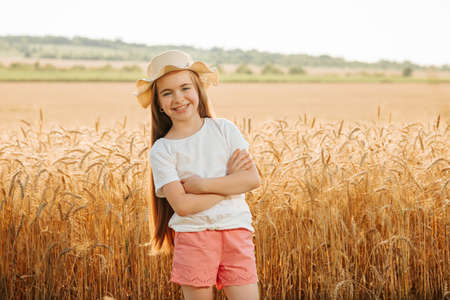 adorable happy little girl with hat in the wheat field, looking at the camera with her hands crossed. family of farmers.の写真素材