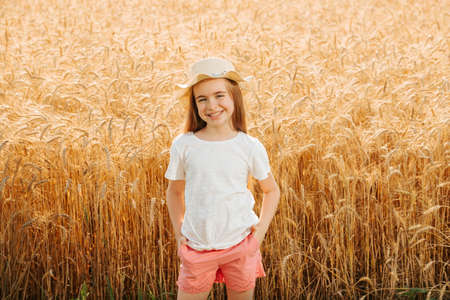 little girl farmers daughter in the wheat field looking at camera and smiles attractively. adorable little child with hat in farmlandの写真素材
