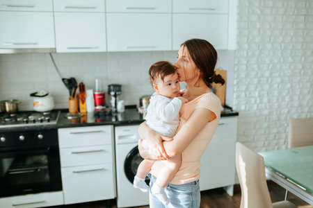 mother embrace baby at home in the kitchen. Child on hands playing laughing and smiling, young beautiful mom and girlの写真素材