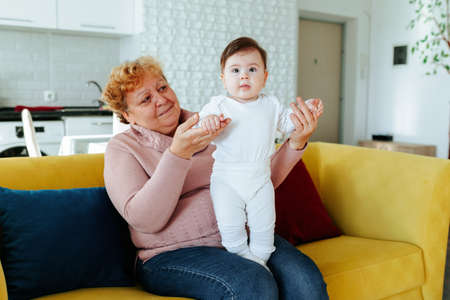 Grandmother with her cute granddaughter baby, smiling, talking, playing. Happy relaxed older adult granny embracing preschool child granddaughter cuddling bonding rest on couch at home.の写真素材