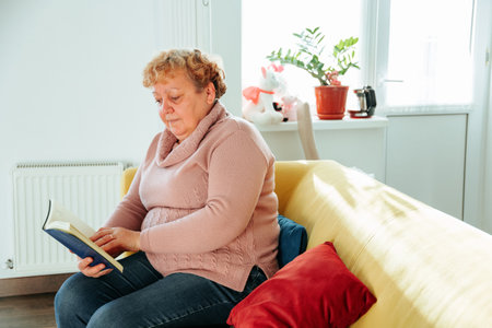 Adult mature fat lady enjoying hobby spending free leisure time with funny modern novel literature alone in living room. elderly woman sitting on yellow sofa in bright living room.の写真素材