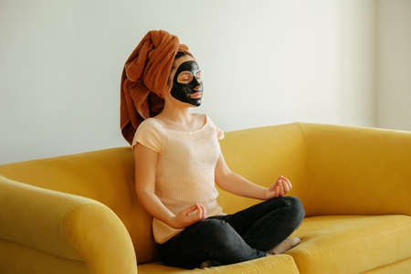 Young woman sitting on a yellow sofa with face beauty mask and doing yoga or meditation. Clean and perfect skin.Yoga poses, cosmetology, hydrationの写真素材