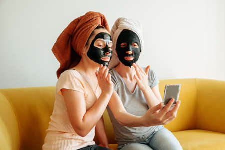 two girls with a towel on their head and a black skin care mask. they look in the mirror and touch their faces with their fingers.の写真素材