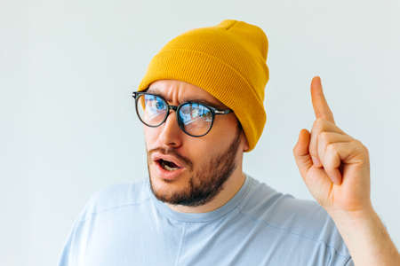 Caucasian handsome man with glasses and a yellow hat pointing up and surprised over isolated white background. copy space. showing and pointing up with finger number oneの写真素材