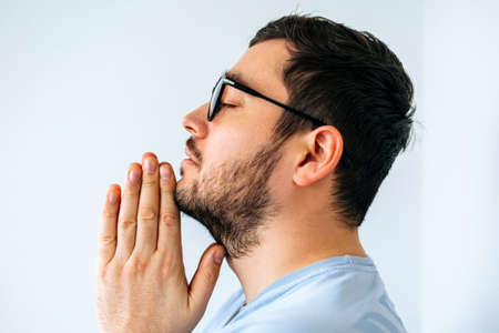 close up of a calm spiritual handsome american guy with glasess praying with closed eyes. side view of a serious peaceful young man with joining hands meditating. Belief conceptの写真素材